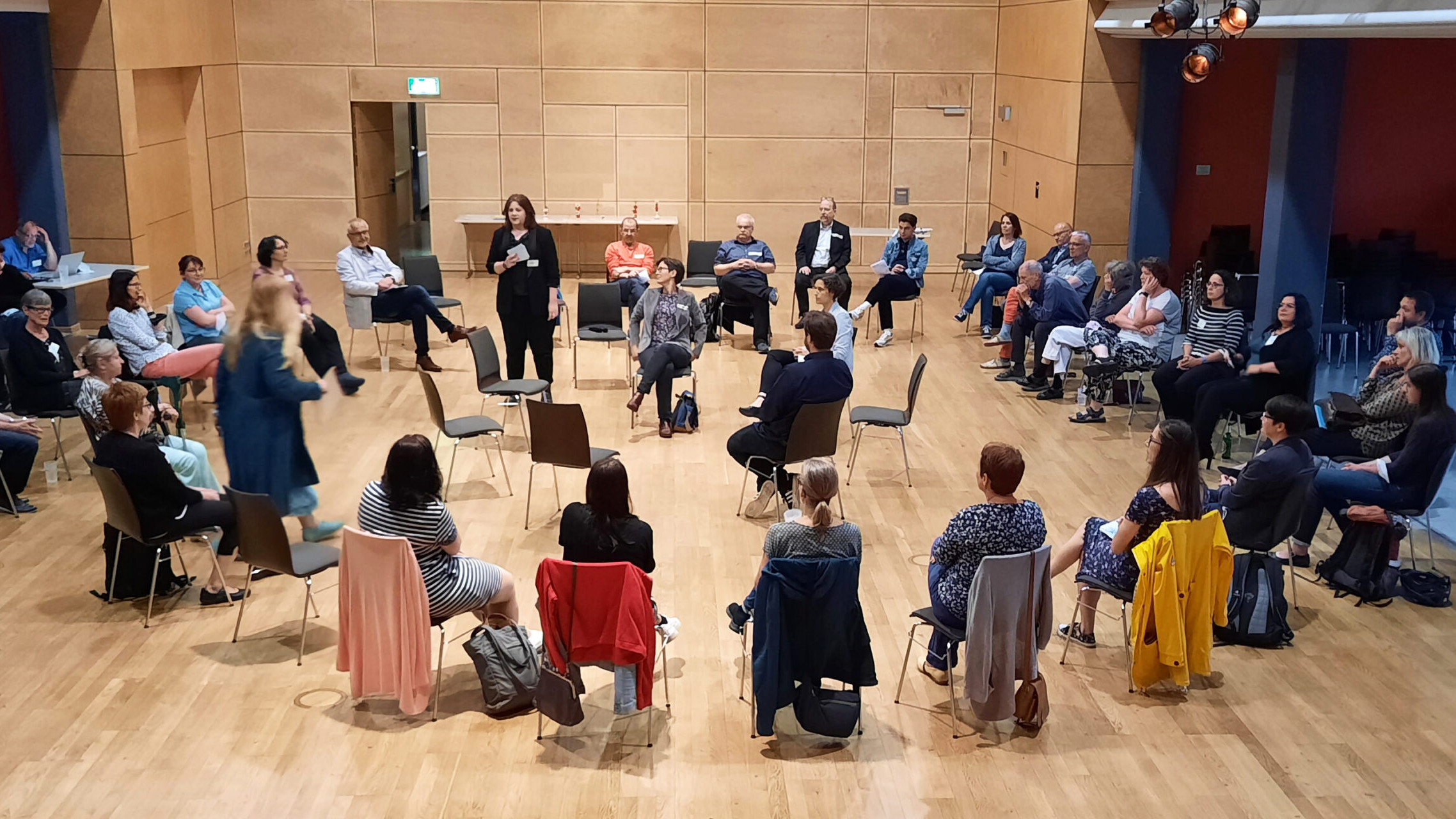 KI generiert: Das Bild zeigt eine Gruppe von Menschen, die in einem Kreis in einem Holzpaneel-Raum sitzen und sich anscheinend an einer Diskussion oder einem Workshop beteiligen. Der Hauptinhalt des Bildes ist eine interaktive Gruppenaktivität oder ein Meeting.