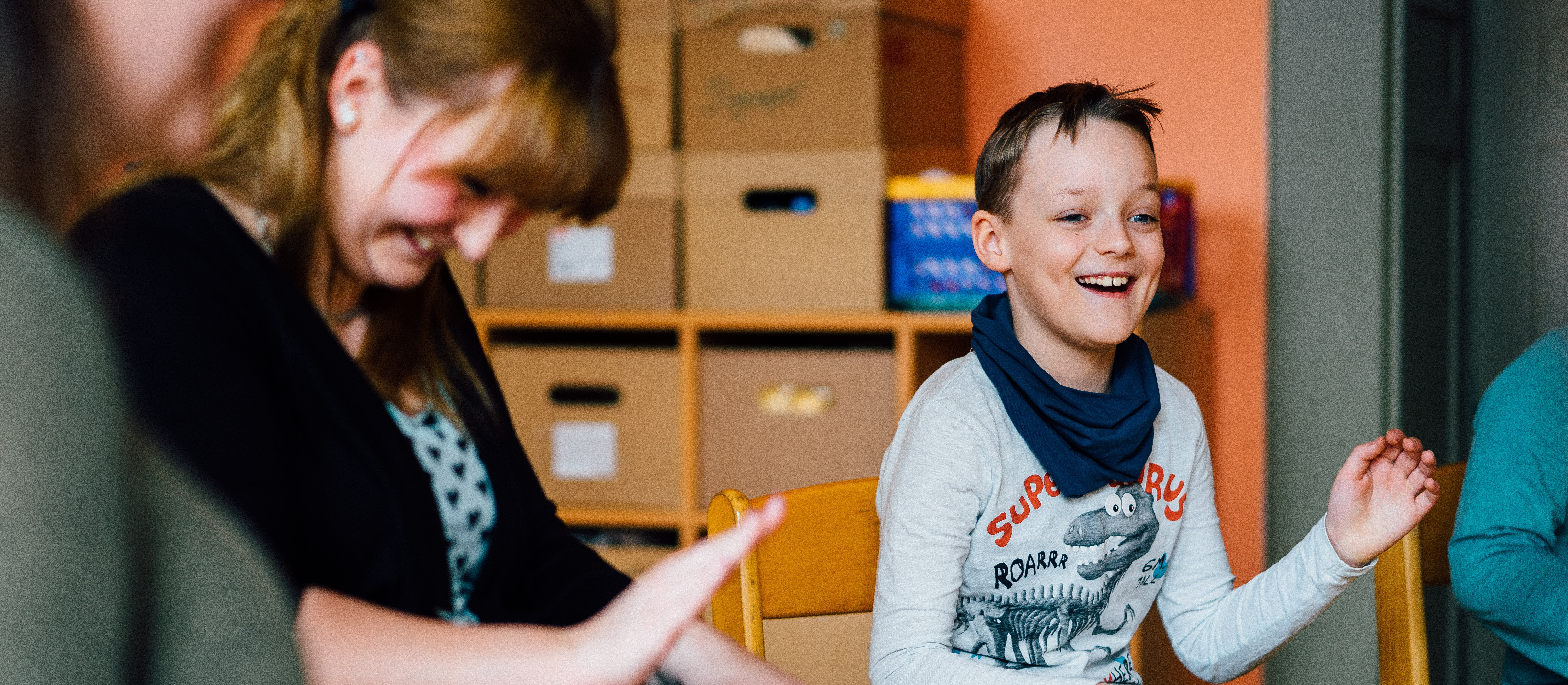 KI generiert: Das Bild zeigt einen lachenden Jungen, der an einem Tisch sitzt und sich freut, während eine Frau neben ihm etwas in den Händen hält. Im Hintergrund sind Regale mit Kartons und Spielzeug zu sehen, was auf eine spielerische oder pädagogische Umgebung hindeutet.