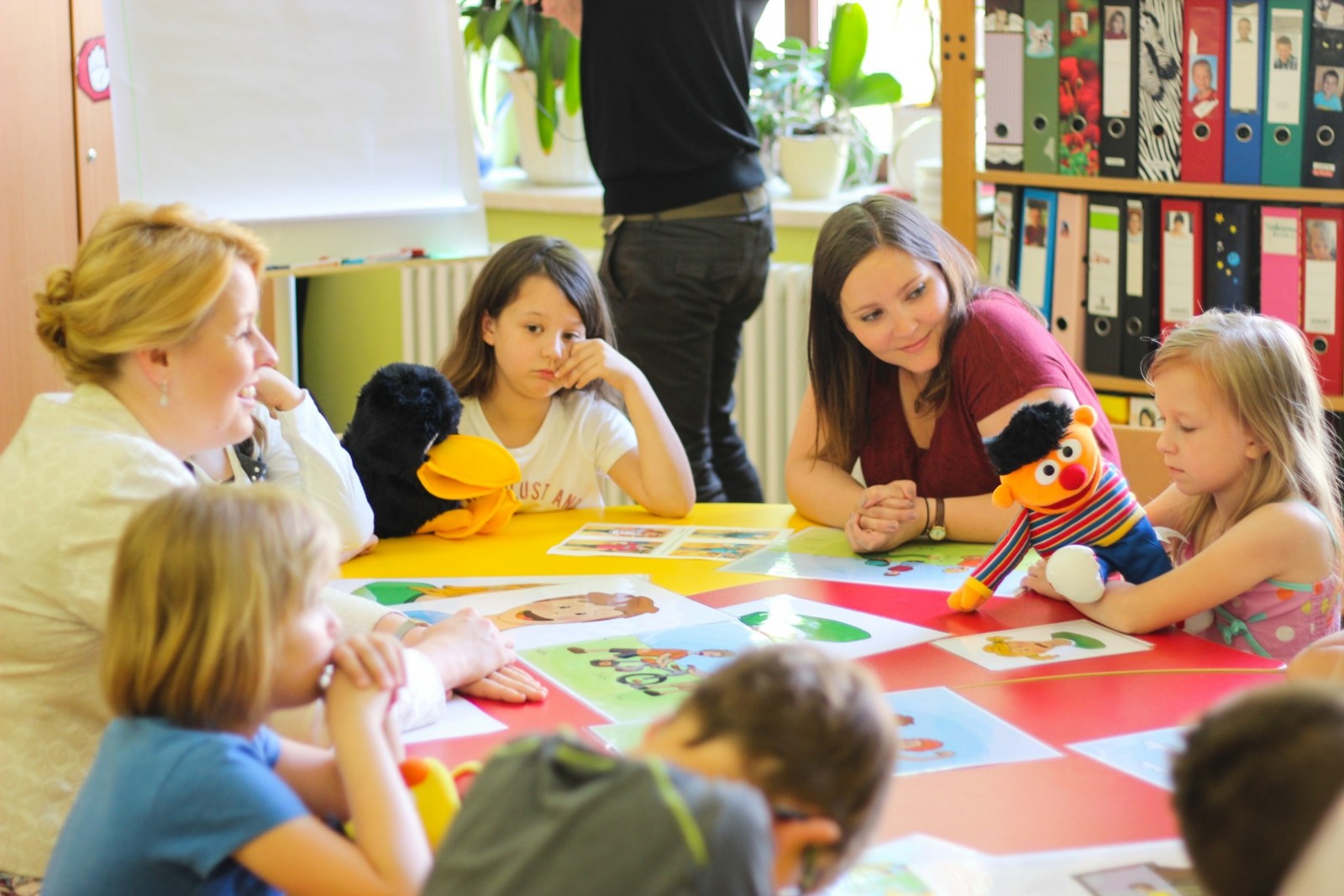 KI generiert: Das Bild zeigt eine Gruppe von Kindern und Erwachsenen, die um einen bunten Tisch sitzen und scheinbar an einer Aktivität oder einem Spiel teilnehmen. Der Hauptinhalt des Bildes ist eine pädagogische oder spielerische Interaktion in einer Kindertagesstätte oder einem Klassenzimmer.