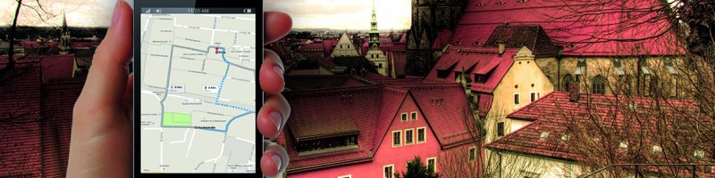KI generiert: Das Bild zeigt eine Hand, die ein Smartphone mit einer Kartenanwendung hält, vor dem Hintergrund einer malerischen Stadtlandschaft mit roten Dächern und historischen Gebäuden. Der Hauptinhalt des Bildes ist die Kombination von moderner Technologie (Smartphone-Navigation) und traditioneller Architektur.