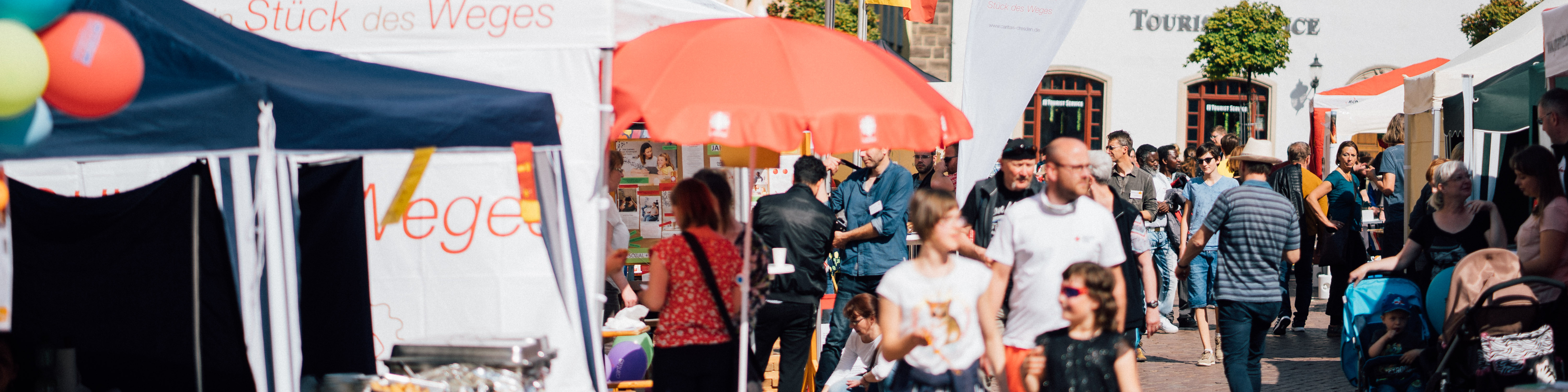 KI generiert: Das Bild zeigt eine belebte Straßenveranstaltung mit mehreren Ständen, darunter ein Stand mit der Aufschrift "Stück des Weges". Menschen schlendern durch die Gassen, betrachten die Angebote und interagieren miteinander.