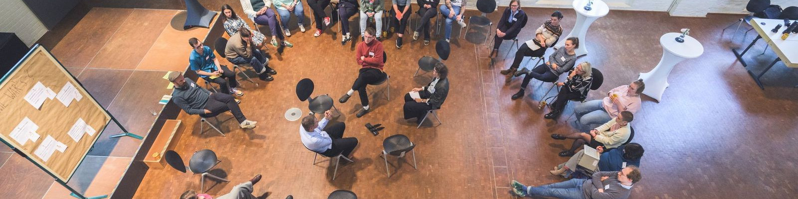 KI generiert: Das Bild zeigt eine Gruppe von Menschen, die in einem Kreis sitzen und sich in einer informellen Besprechung oder Diskussion befinden. Der Hauptinhalt des Bildes ist eine interaktive Sitzung oder ein Workshop in einem hellen, modernen Innenraum.