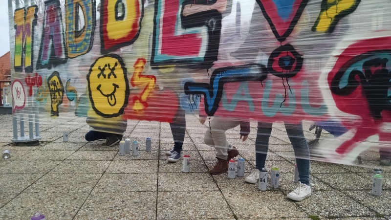 KI generiert: Das Bild zeigt mehrere Personen, die an einer großen Graffiti-Wand arbeiten, auf der bunte Buchstaben und Symbole zu sehen sind. Im Vordergrund liegen mehrere Spraydosen auf dem Boden.