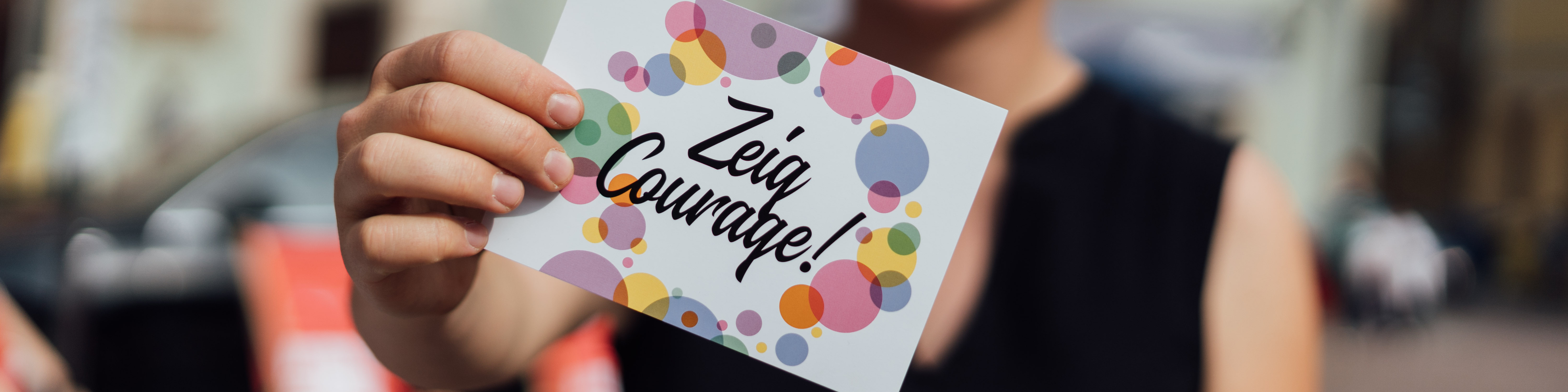 KI generiert: Das Bild zeigt eine Person, die ein weißes Blatt Papier mit der Aufschrift "Zeiß Courage!" und bunten Kreisen im Hintergrund hält. Der Hauptinhalt des Bildes ist die ermutigende Botschaft auf dem Blatt Papier.
