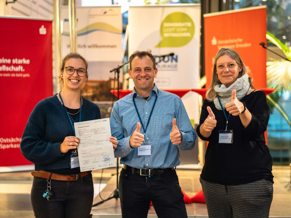 KI generiert: Das Bild zeigt zwei Personen, die lächelnd vor mehreren Roll-up-Bannern stehen und jeweils ein Dokument in der Hand halten. Die Banner beinhalten Begrüßungen und Informationen zu verschiedenen Themen wie Demokratie, Zivilcourage und regionalen Angeboten.