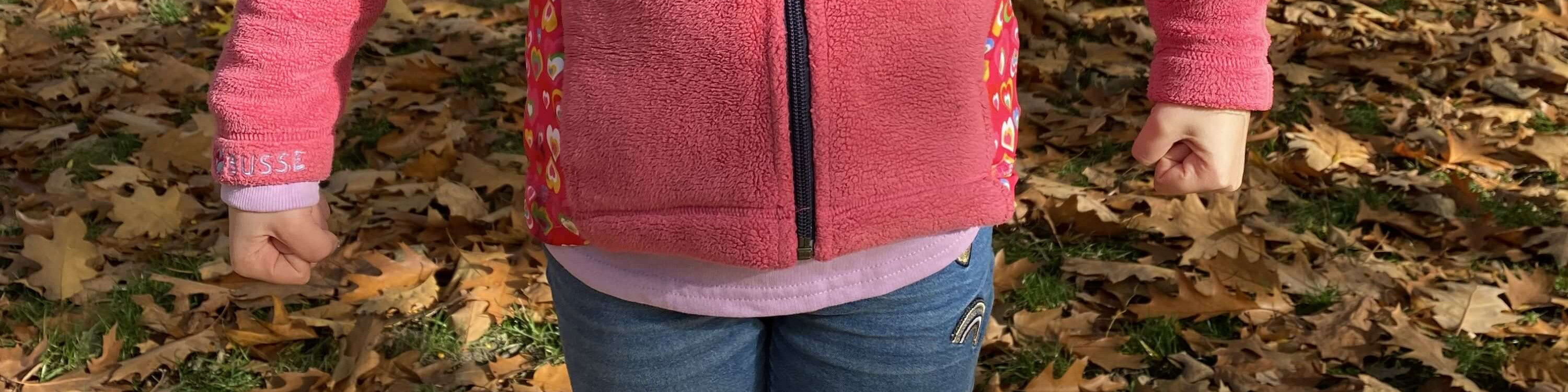 KI generiert: Das Bild zeigt eine Person von hinten, die eine pinkfarbene Jacke mit bunten Mustern auf den Ärmeln und eine helle Bluse trägt, während sie auf einem Boden voller herbstlicher Blätter steht. Der Hauptinhalt des Bildes ist die Kleidung der Person und die herbstliche Umgebung.