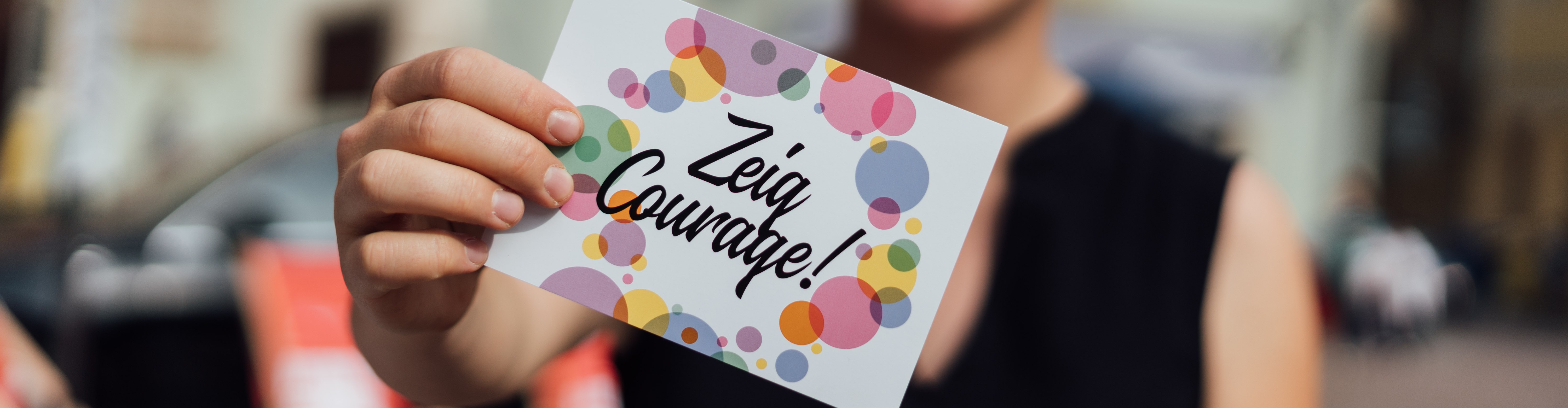 KI generiert: Eine Person hält eine Karte mit der Aufschrift "Zeig Courage!" in der Hand. Die Karte ist bunt mit vielen Kreisen gestaltet.