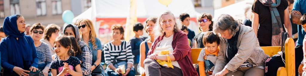 KI generiert: Das Bild zeigt eine Gruppe von Menschen, die auf Bänken sitzen und einer Veranstaltung im Freien zuhören. Der Hauptinhalt ist eine Gemeinschaftsveranstaltung, bei der Menschen verschiedenen Alters zusammenkommen.