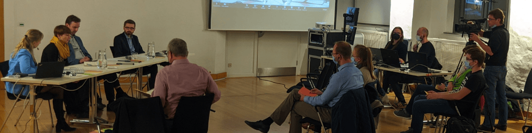 KI generiert: Das Bild zeigt eine Gruppe von Personen in einem Seminarraum, die an langen Tischen sitzen und sich unterhalten oder einer Präsentation folgen. Im Vordergrund sind einige Teilnehmer zu sehen, während im Hintergrund eine Leinwand mit einer Präsentation und ein Kameramann, der die Veranstaltung aufzeichnet, sichtbar sind.
