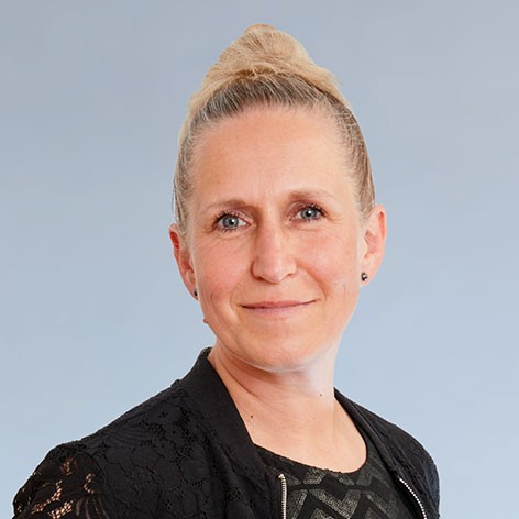 KI generiert: Das Bild zeigt eine lächelnde Frau mit hellen Haaren, die zu einem Dutt gebunden sind. Sie trägt ein schwarzes, gemustertes Oberteil und hat einen neutralen Hintergrund.