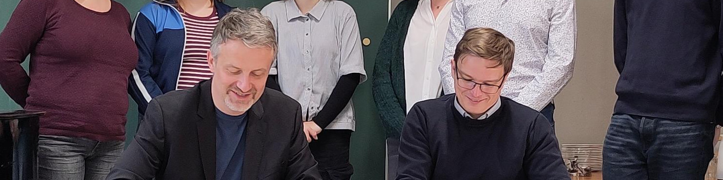 KI generiert: Das Bild zeigt zwei Männer, die an einem Tisch sitzen und Dokumente unterschreiben, während eine Gruppe von Personen im Hintergrund steht und zuschaut. Der Hauptinhalt des Bildes ist eine offizielle Unterzeichnung oder ein Vertragsabschluss.