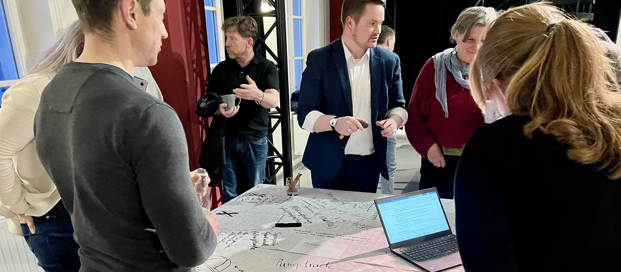 KI generiert: Das Bild zeigt eine Gruppe von Personen, die sich um einen Tisch versammelt haben und eine Diskussion führen. Auf dem Tisch liegen verschiedene Unterlagen und ein Laptop, was auf eine Besprechung oder ein Meeting hindeutet.