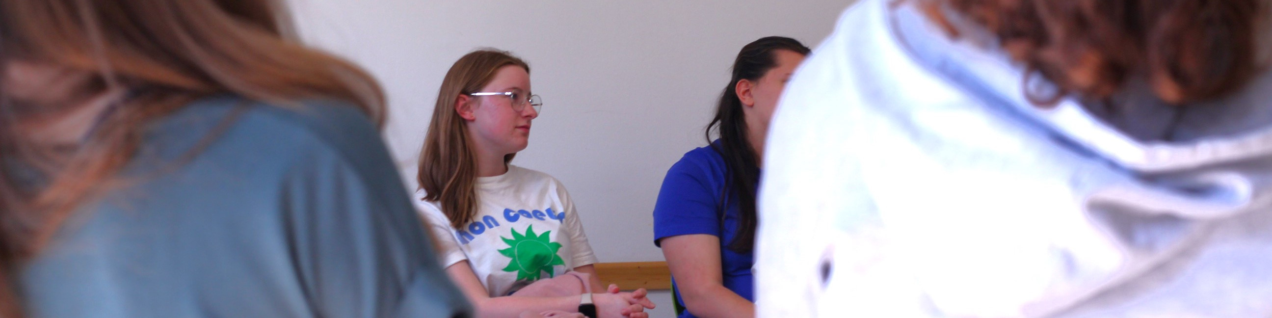KI generiert: Das Bild zeigt eine Gruppe von Personen, die in einem Raum stehen und sich unterhalten. Der Hauptinhalt ist eine Person mit Brille und einem T-Shirt mit einem Aufdruck, die im Mittelpunkt der Gruppe steht und sich mit anderen unterhält.