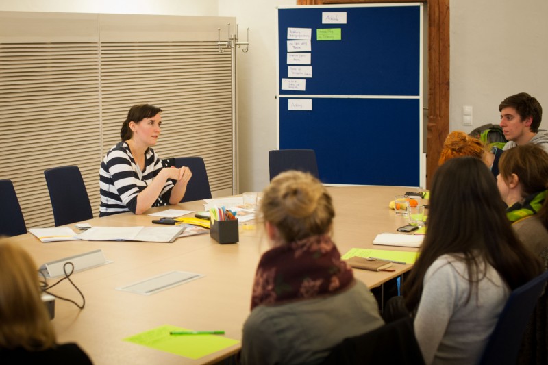 KI generiert: Das Bild zeigt eine Besprechung oder ein Seminar in einem Konferenzraum, bei dem eine Frau an einem Tisch mit mehreren Teilnehmern spricht. Im Hintergrund hängt ein blaues Whiteboard mit Notizen und Post-its.