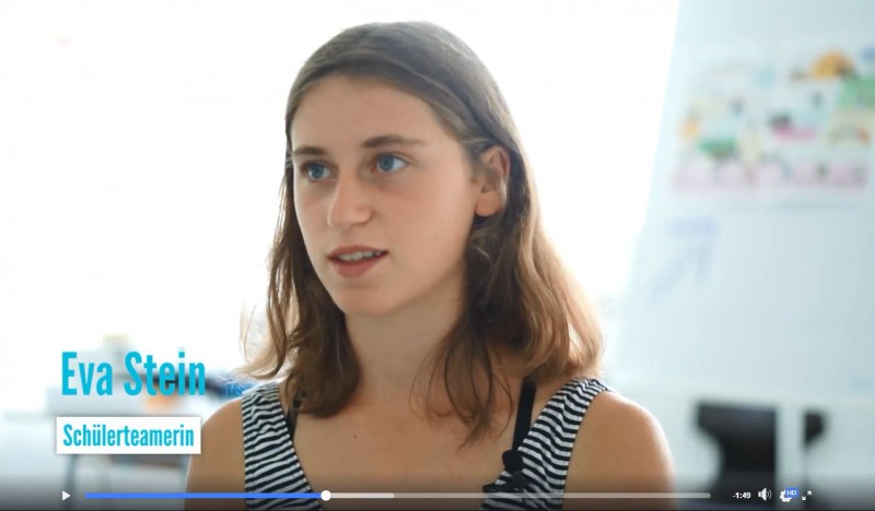 KI generiert: Das Bild zeigt eine junge Frau namens Eva Stein, die als Schülerteamerin bezeichnet wird. Sie scheint in einem Interview oder einer Diskussion zu sprechen, während im Hintergrund eine Tafel mit Notizen sichtbar ist.
