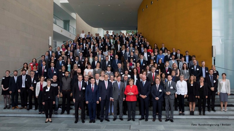 KI generiert: Das Bild zeigt eine große Gruppe von Menschen, die auf einer Treppe posieren, wobei viele von ihnen Namensschilder tragen. Im Mittelpunkt steht eine Frau in einem roten Blazer, die von mehreren Personen umgeben ist.