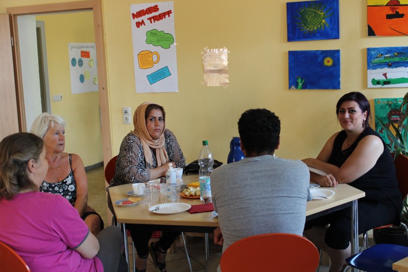 KI generiert: Das Bild zeigt eine Gruppe von Personen, die an einem Tisch sitzen und sich unterhalten, möglicherweise in einer informellen Besprechung oder einem Workshop. Der Raum ist mit bunten Postern und Kinderzeichnungen an den Wänden dekoriert.