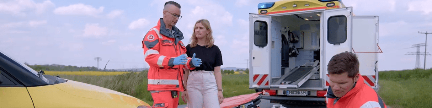 KI generiert: Das Bild zeigt eine Notfallsituation, bei der zwei Rettungssanitäter in orangefarbener Schutzkleidung und eine Frau neben einem Unfallfahrzeug und einem offenen Rettungswagen stehen. Der Hauptinhalt des Bildes ist die Darstellung einer Rettungsaktion nach einem Verkehrsunfall.