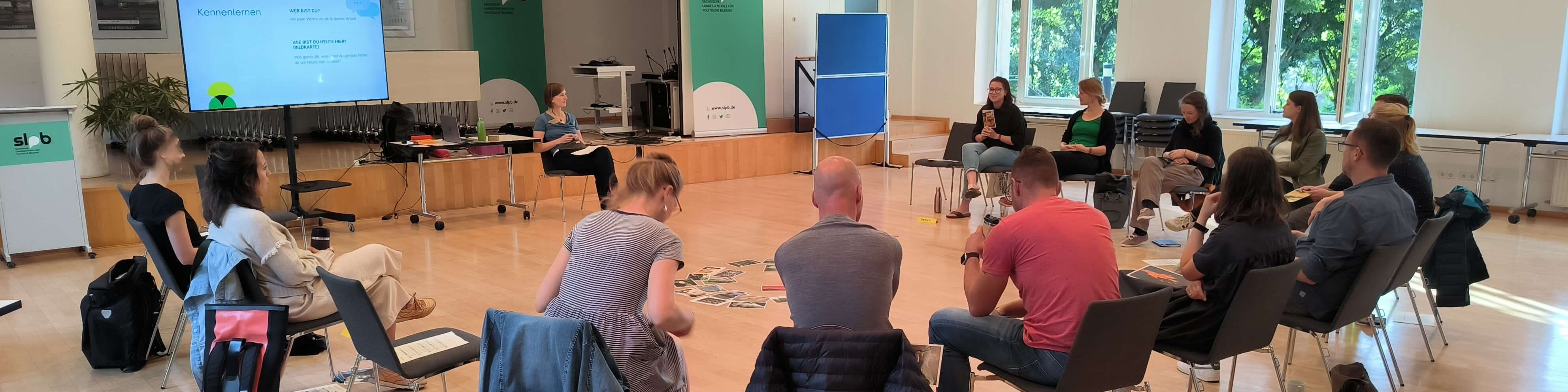 KI generiert: Das Bild zeigt eine Gruppe von Personen, die in einem hellen Raum mit großen Fenstern an einem Workshop oder einer Besprechung teilnehmen. Im Vordergrund sitzen einige Teilnehmer auf Stühlen, während andere auf dem Boden sitzen und Notizen machen oder zuhören.
