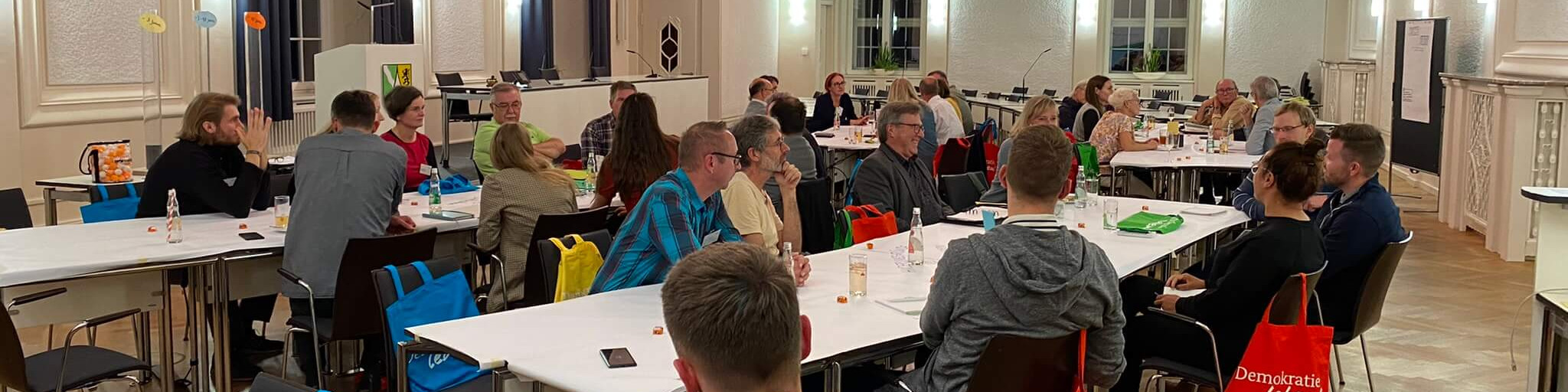 KI generiert: Das Bild zeigt eine Gruppe von Menschen, die in einem hellen Raum mit großen Fenstern an langen Tischen sitzen und sich unterhalten. Der Hauptinhalt des Bildes ist eine Versammlung oder ein Treffen, möglicherweise eine Konferenz oder ein Workshop.