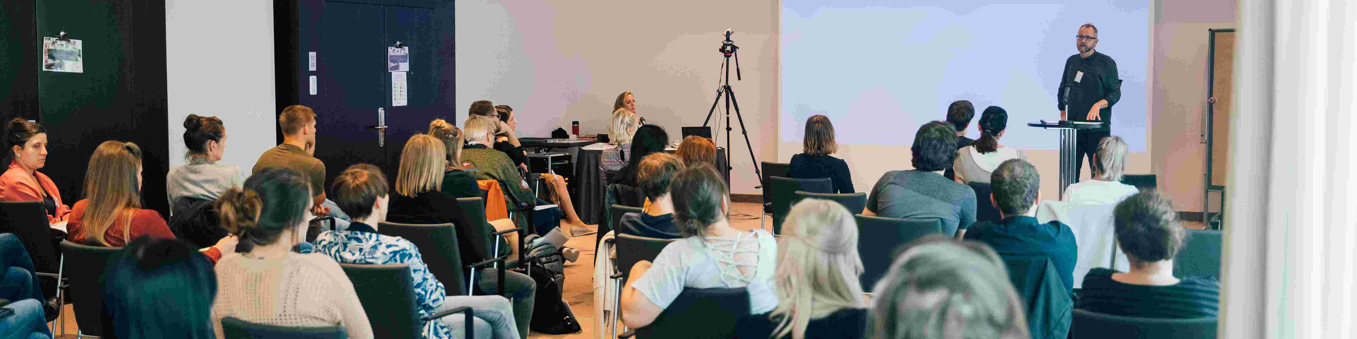 KI generiert: Das Bild zeigt eine Gruppe von Personen in einem Seminarraum, wobei ein Mann vorne steht und zu den Teilnehmern spricht. Im Hintergrund sitzt eine Frau an einem Tisch mit einem Mikrofon, während die Zuhörer auf Stühlen Platz genommen haben.