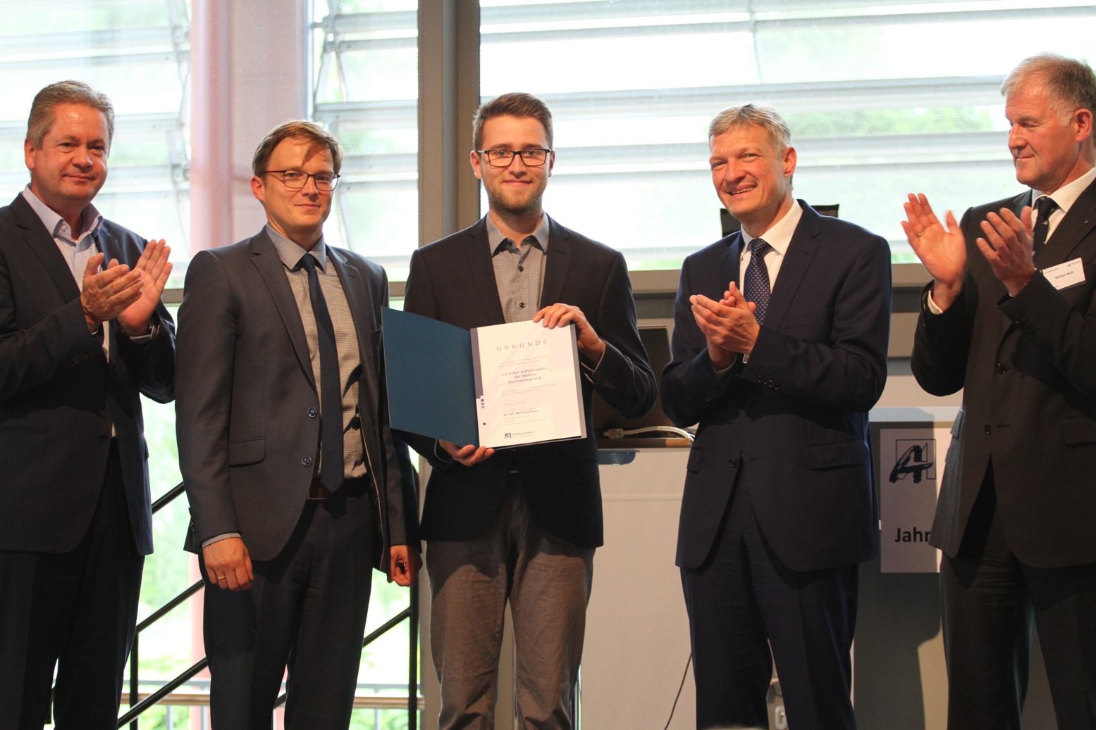 KI generiert: Das Bild zeigt eine Gruppe von fünf Männern in formeller Kleidung, wobei einer von ihnen eine Urkunde oder ein Buch in den Händen hält. Die anderen vier applaudieren, was darauf hindeutet, dass eine Auszeichnung oder Anerkennung stattfindet.