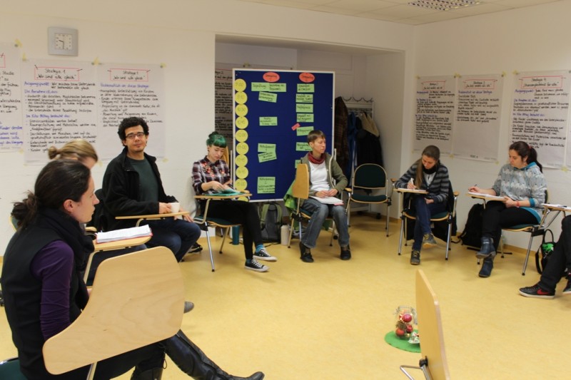 KI generiert: Das Bild zeigt eine Gruppe von Personen, die in einem Klassenzimmer oder Seminarraum sitzen und scheinbar an einer Diskussion oder einem Workshop teilnehmen. Im Hintergrund sind verschiedene Poster und Notizen an den Wänden zu sehen, die auf eine interaktive Lernumgebung hindeuten.