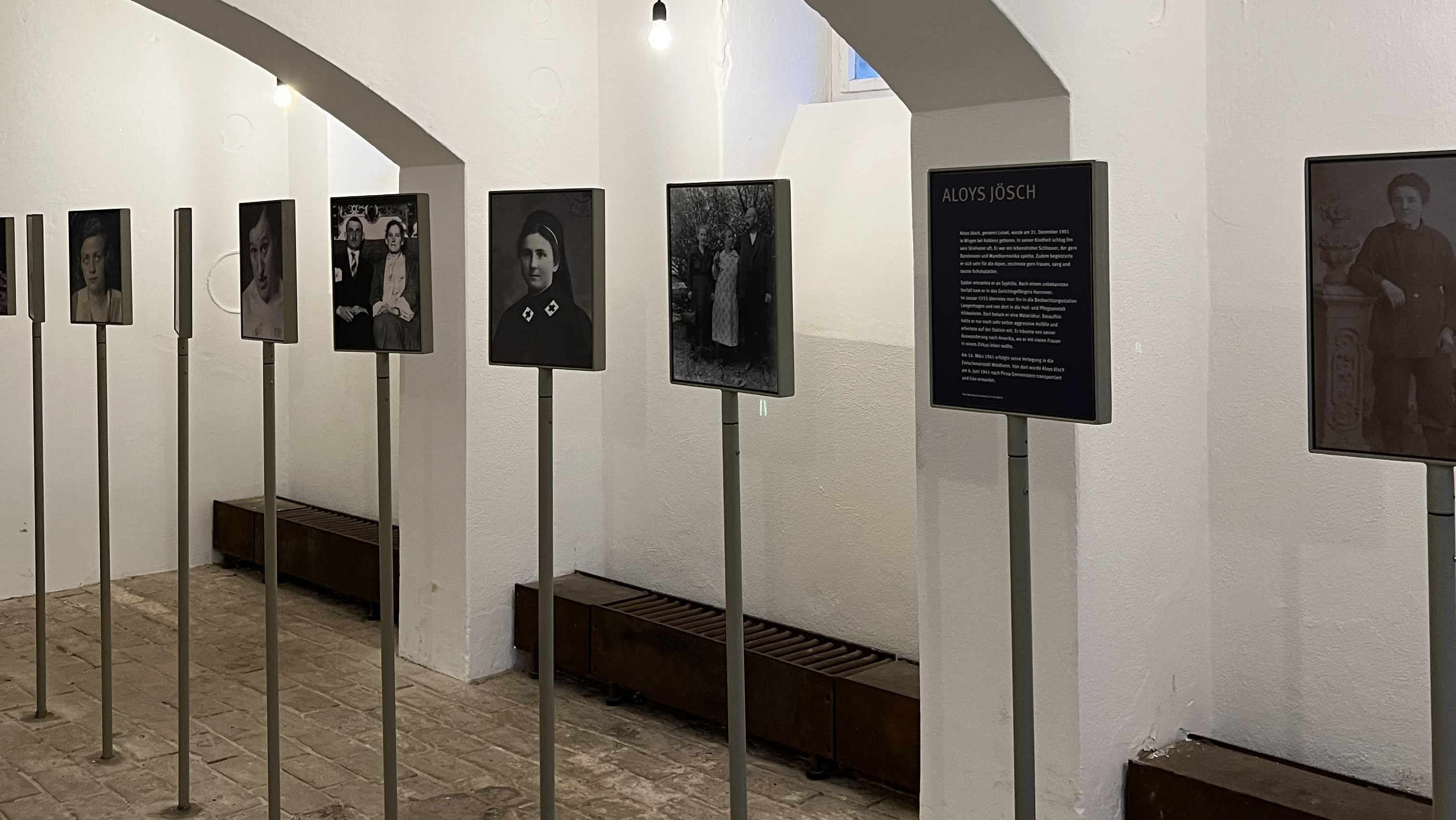KI generiert: Das Bild zeigt eine Ausstellung in einem Museum oder einer Galerie mit mehreren schwarz-weißen Fotografien, die an dünnen Metallstangen befestigt sind. Der Hauptinhalt der Ausstellung scheint historische Porträts und eine Informationsplatte über Aloys Jösch zu sein.