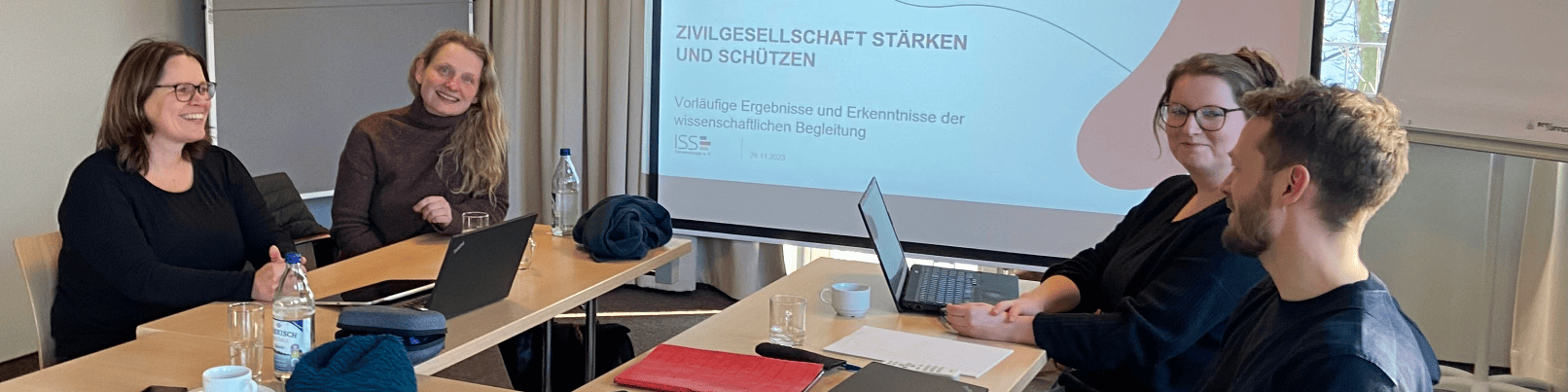 KI generiert: Das Bild zeigt eine Gruppe von vier Personen, die an einem Tisch sitzen und an Laptops arbeiten, während eine Präsentation im Hintergrund auf einem Bildschirm läuft. Der Hauptinhalt des Bildes scheint eine Arbeitsbesprechung oder ein Workshop zu sein, bei dem die Teilnehmer gemeinsam an einem Projekt arbeiten.