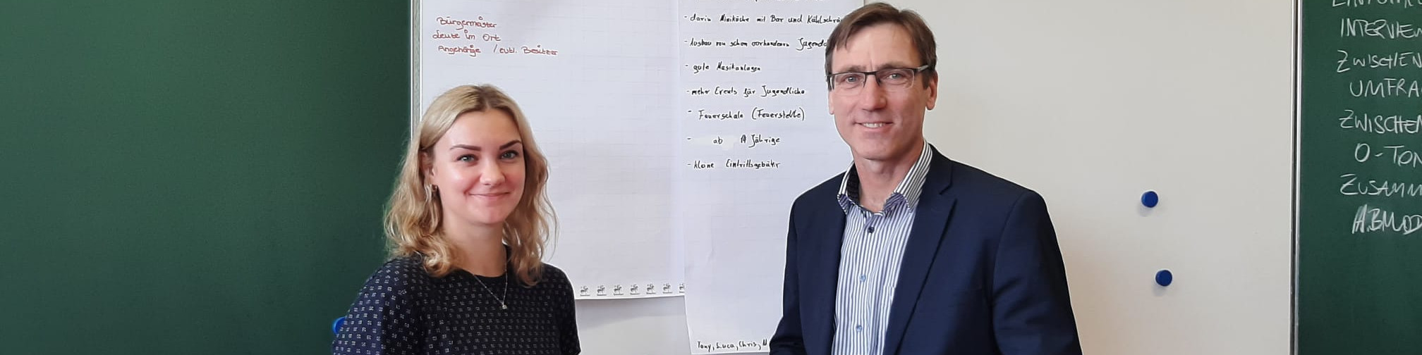 KI generiert: Das Bild zeigt zwei Personen, eine Frau und einen Mann, die vor einem Whiteboard mit handschriftlichen Notizen stehen. Der Hauptinhalt des Bildes ist eine scheinbar akademische oder geschäftliche Besprechung oder Präsentation.