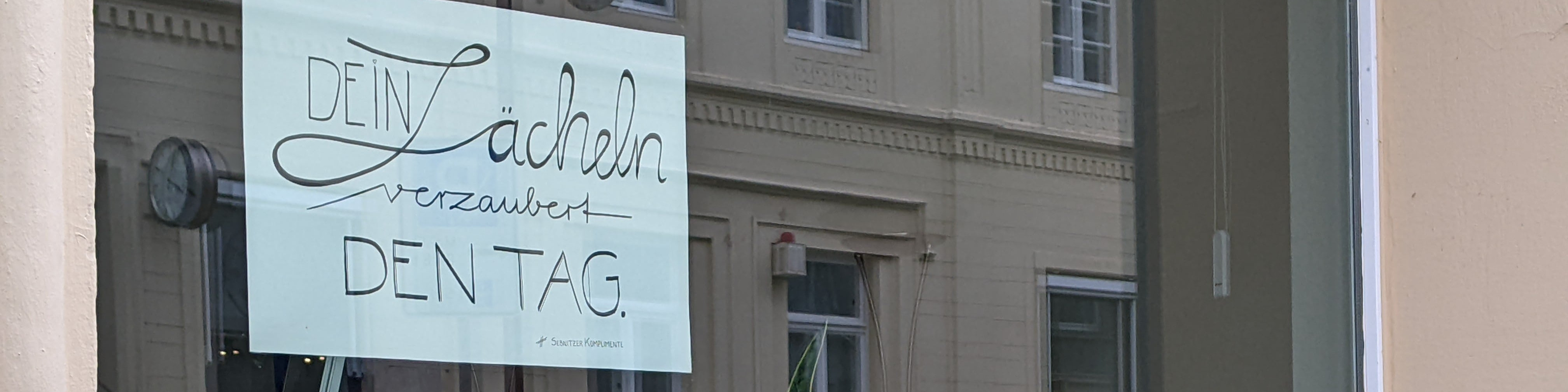 KI generiert: Das Bild zeigt ein Schaufenster mit einem handgeschriebenen Zettel, auf dem steht: "Dein Lächeln verzaubert den Tag." Im Hintergrund sind die Fassade eines Gebäudes und ein Teil des Innenraums des Ladens zu sehen. Der Hauptinhalt des Bildes ist die positive Botschaft auf dem Zettel.