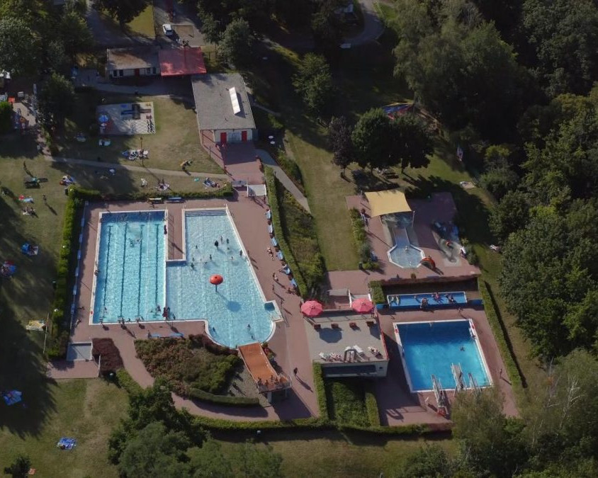KI generiert: Das Bild zeigt eine Luftaufnahme eines Schwimmbads mit mehreren Becken, umgeben von Grünflächen und Gebäuden. Der Hauptinhalt des Bildes ist das große, belebte Freibad mit verschiedenen Wasserbecken und Liegeflächen.
