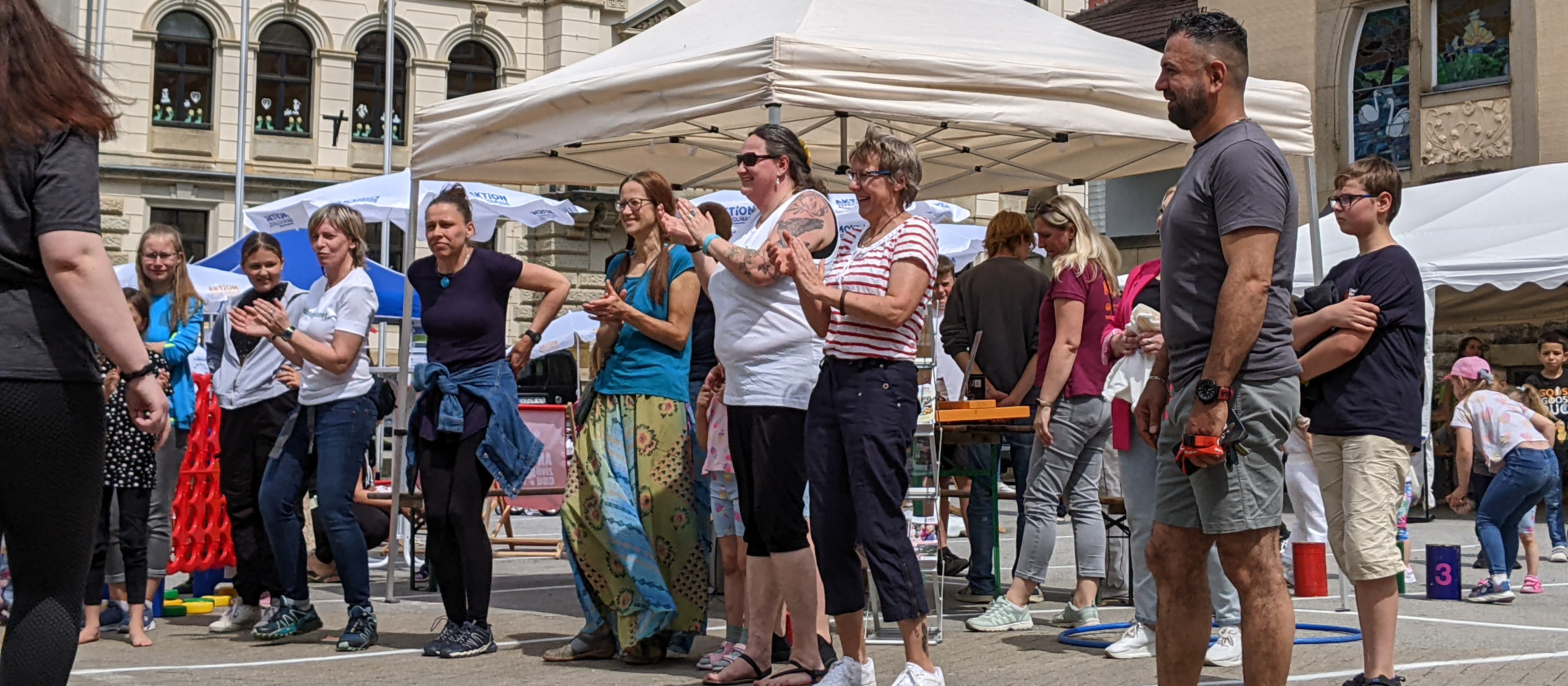 KI generiert: Das Bild zeigt eine Gruppe von Menschen, die sich auf einem öffentlichen Platz versammelt haben, möglicherweise bei einem Event oder einer Feier. Im Vordergrund stehen mehrere Personen, die sich unterhalten oder zuschauen, während im Hintergrund weitere Menschen und ein Zelt zu sehen sind.