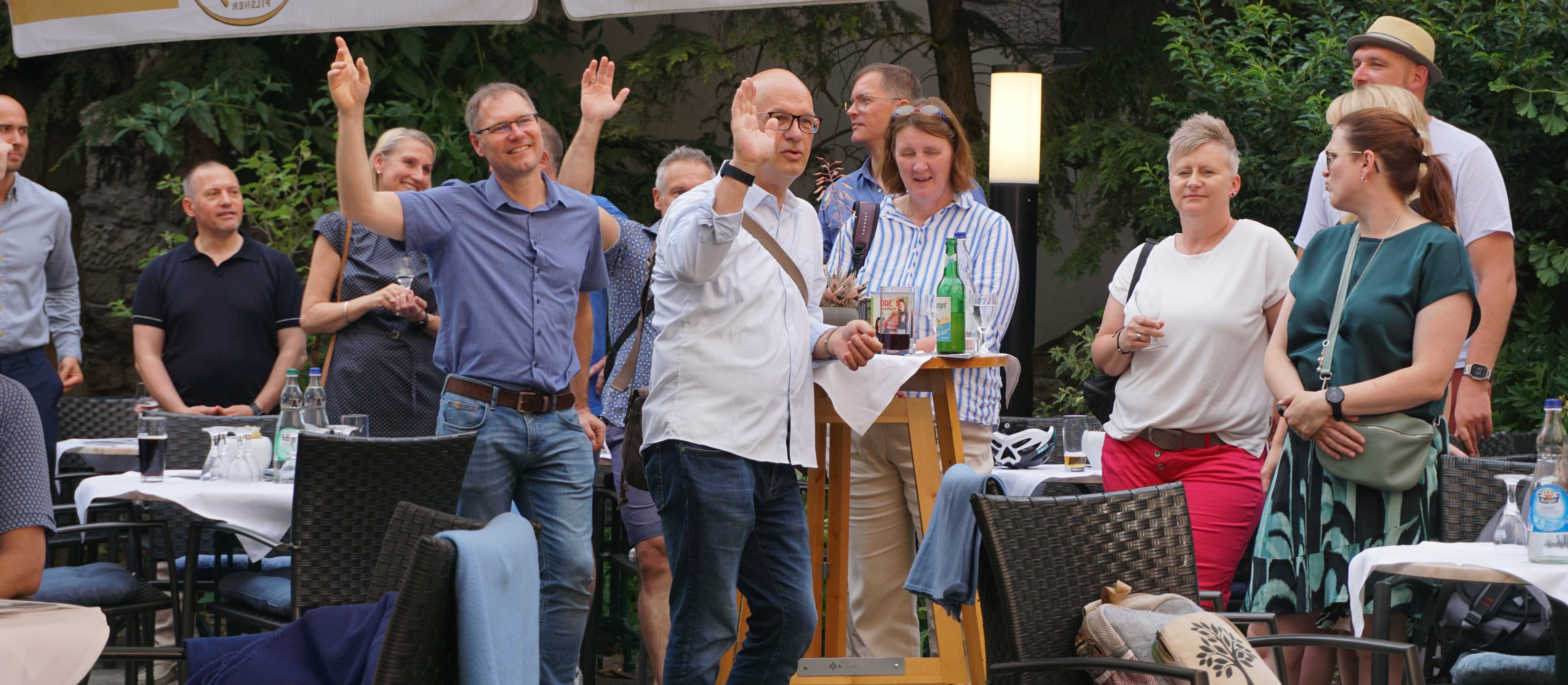 KI generiert: Das Bild zeigt eine Gruppe von Menschen, die sich in einem Freiluftbereich mit Tischen und Stühlen versammelt haben. Zwei Männer im Vordergrund scheinen eine interaktive oder spielerische Aktivität durchzuführen, während die anderen zuschauen und sich unterhalten.