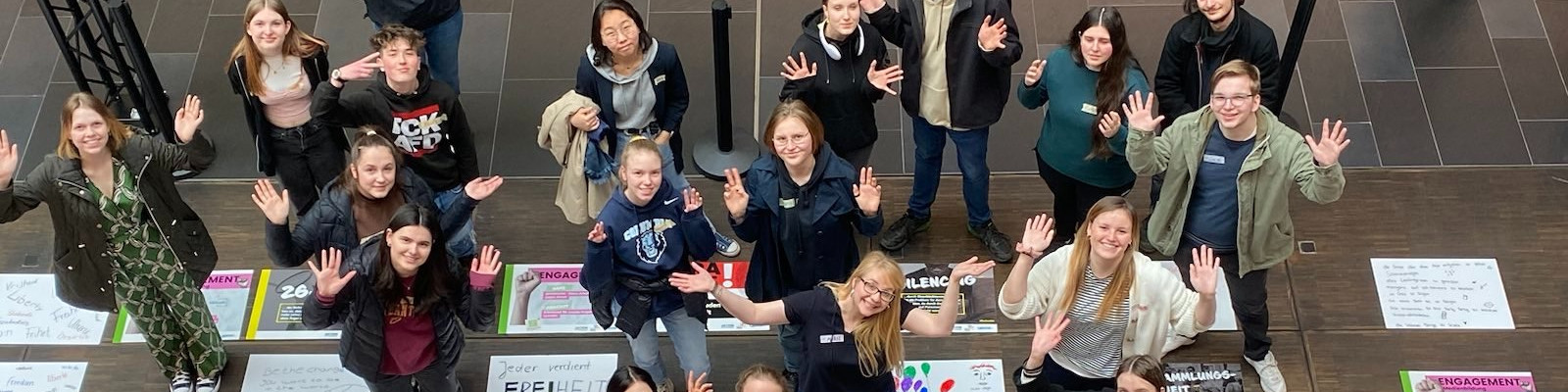 KI generiert: Das Bild zeigt eine Gruppe von Personen, die auf dem Boden sitzen und stehend in einem Innenraum posieren, wobei viele von ihnen mit den Händen winken. Der Hauptinhalt des Bildes ist eine fröhliche und entspannte Gruppensituation, möglicherweise eine Schulklasse oder eine Gruppe von Teilnehmern einer Veranstaltung.