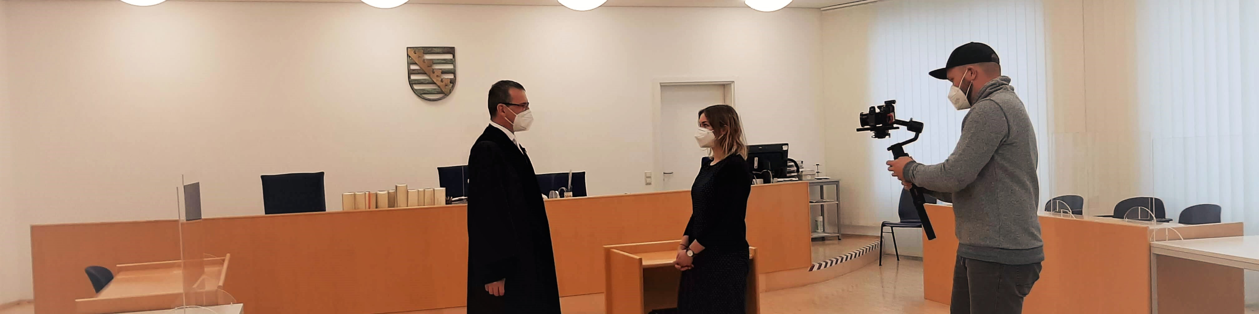 KI generiert: Das Bild zeigt einen Gerichtssaal, in dem eine Person in Robe und eine weitere Person vor einem Richtertisch stehen, während eine dritte Person mit einer Kamera die Szene filmt. Der Hauptinhalt des Bildes ist eine gerichtliche Verhandlung oder ein gerichtliches Verfahren, das aufgezeichnet wird.