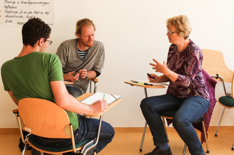 KI generiert: Das Bild zeigt drei Personen, die in einem Raum an Tischen sitzen und sich in einer Diskussion oder einem Meeting befinden. Der Hauptinhalt des Bildes ist eine Gruppeninteraktion, bei der Notizen gemacht und Ideen ausgetauscht werden.