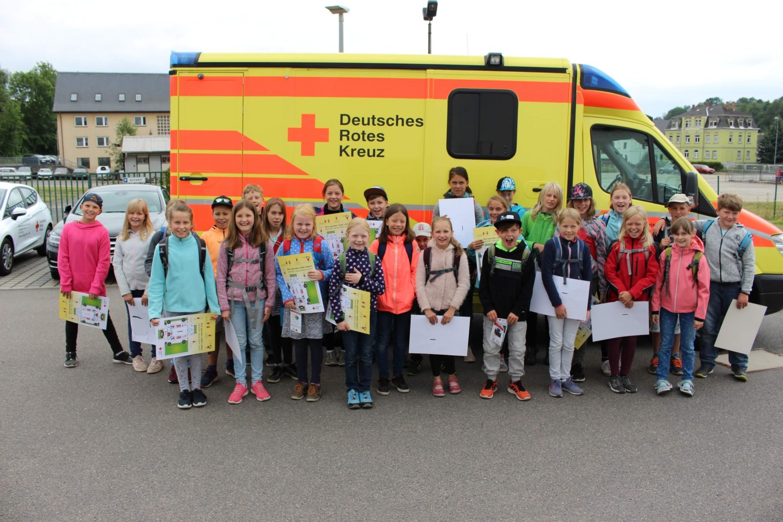 KI generiert: Das Bild zeigt eine Gruppe von Kindern, die vor einem Rettungswagen des Deutschen Roten Kreuzes (DRK) stehen und stolz ihre Zertifikate oder Urkunden präsentieren. Der Hauptinhalt des Bildes ist die feierliche Übergabe von Auszeichnungen, möglicherweise im Rahmen einer Erste-Hilfe-Ausbildung oder eines ähnlichen Programms.
