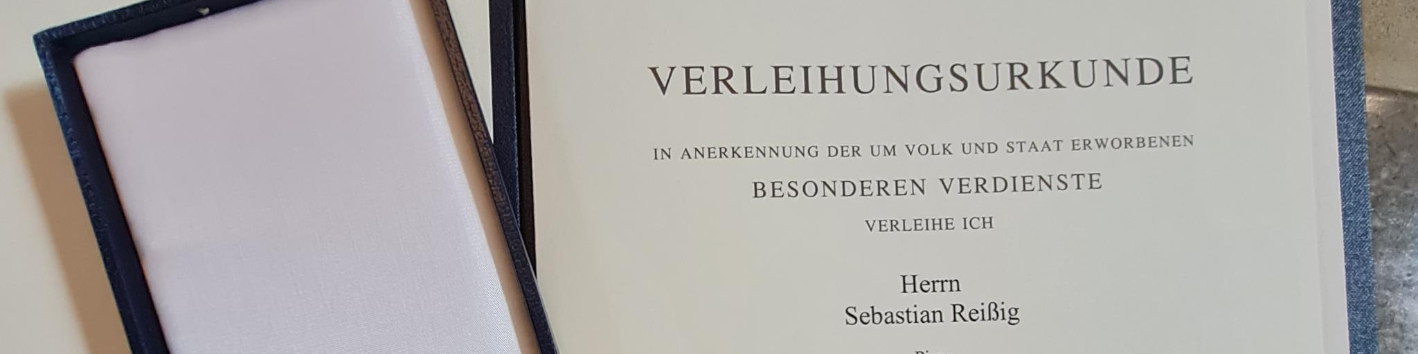 KI generiert: Das Bild zeigt eine Verleihungsurkunde in einem offenen, dunklen Etui. Die Urkunde ist an Herrn Sebastian Reißig für besondere Verdienste verliehen worden.
