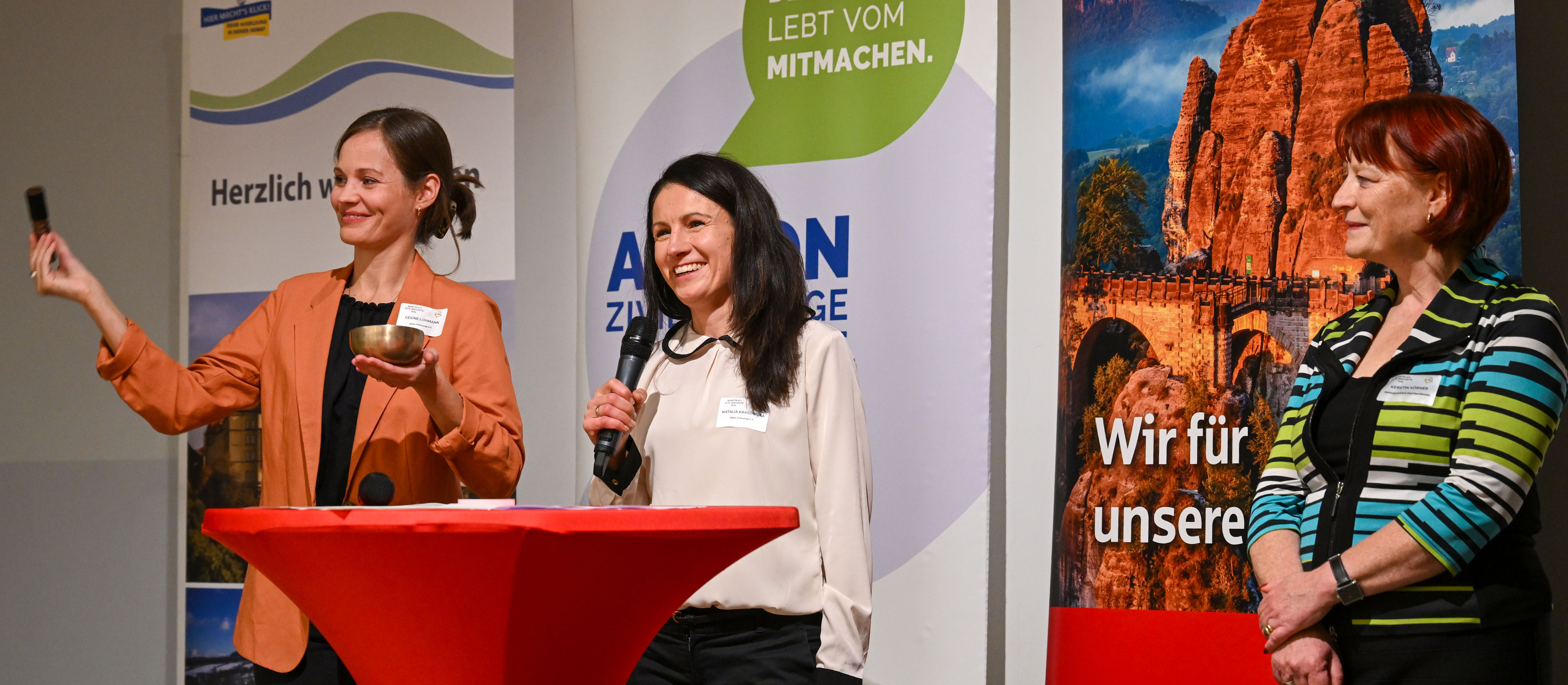 KI generiert: Das Bild zeigt drei Frauen auf einer Bühne, wobei eine Frau an einem Rednerpult steht und spricht, während eine andere Frau daneben steht und eine dritte Frau zuhört. Im Hintergrund sind Banner mit verschiedenen Aufschriften und Bildern zu sehen.