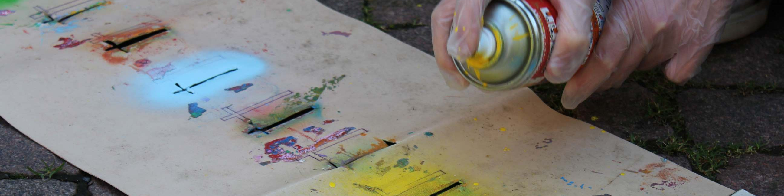 KI generiert: Das Bild zeigt eine Person, die mit einer Spraydose auf einem großen, auf dem Boden liegenden Blatt Papier Graffiti sprüht. Der Hauptinhalt des Bildes ist das Erstellen von Street Art oder Graffiti auf Papier.