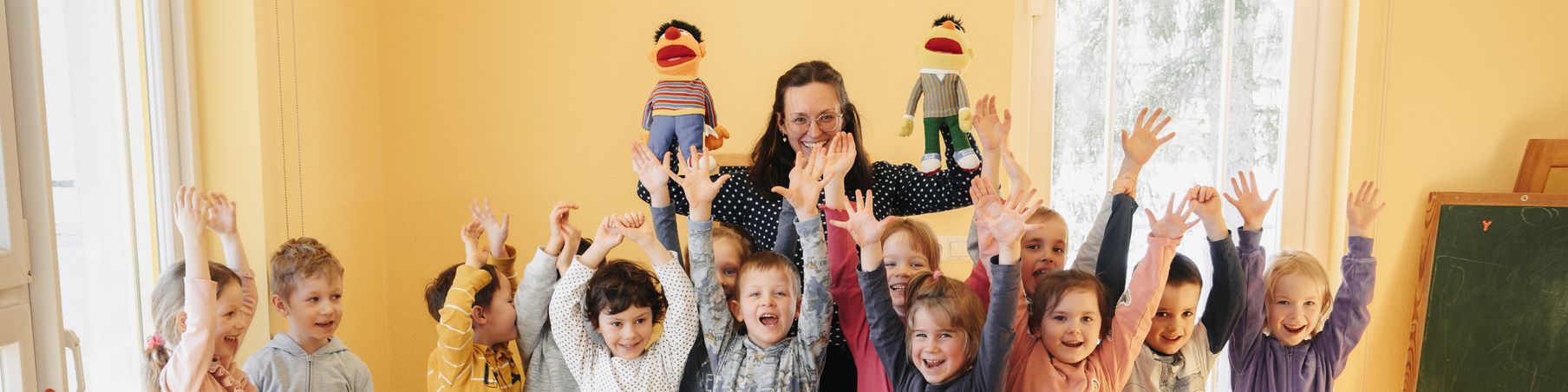 KI generiert: Das Bild zeigt eine Gruppe fröhlicher Kinder, die gemeinsam mit einer Erwachsenen in einem hellen Raum stehen und ihre Hände in die Luft strecken. Im Hintergrund sind zwei Puppen an der Wand zu sehen.