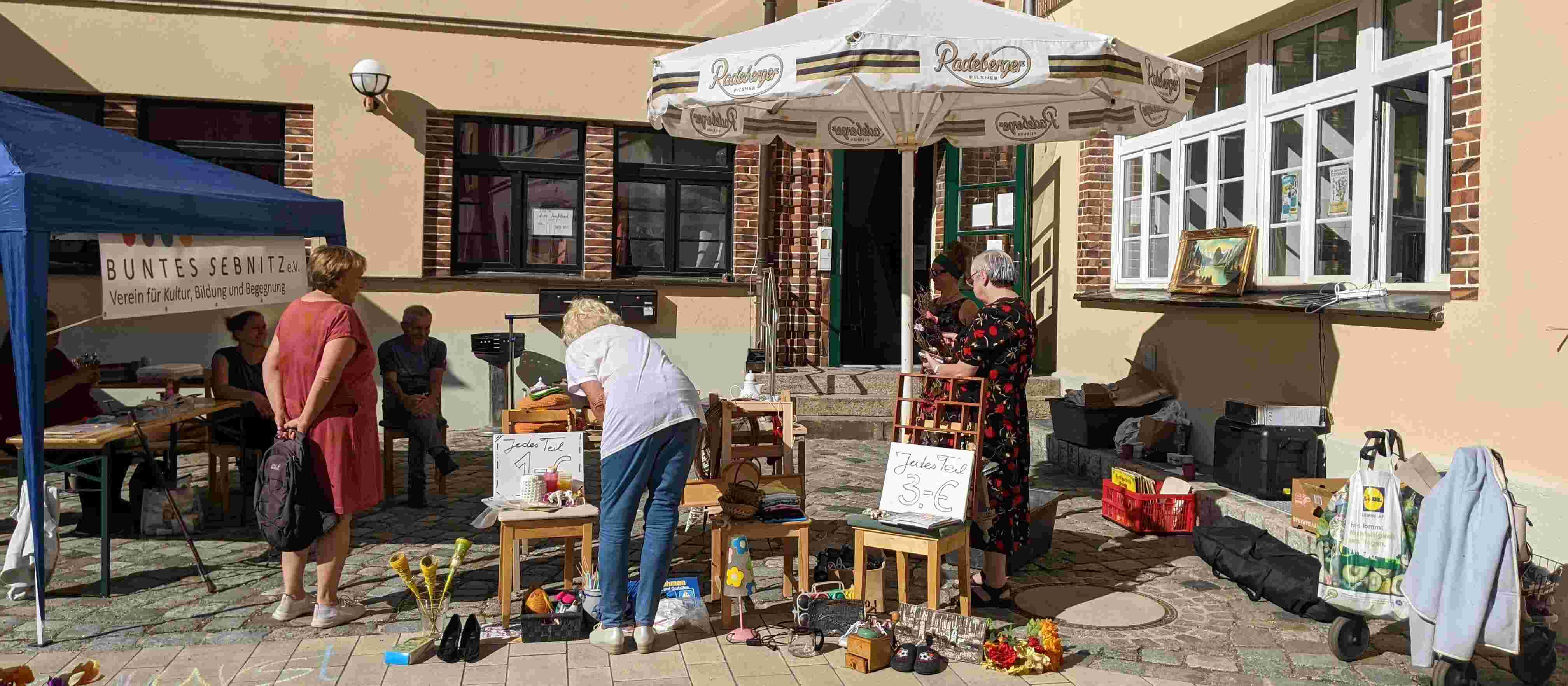 KI generiert: Das Bild zeigt eine kleine Straßenveranstaltung oder einen Flohmarkt vor einem Gebäude mit mehreren Ständen und Tischen, auf denen verschiedene Gegenstände wie Schuhe, Bilder und Haushaltswaren ausgestellt sind. Menschen durchstöbern die angebotenen Artikel und unterhalten sich.