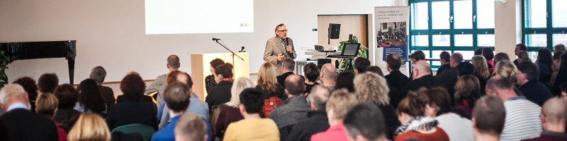 KI generiert: Das Bild zeigt eine Versammlung oder Konferenz mit vielen Teilnehmern, die einem Redner zuhören, der an einem Pult steht. Der Hauptinhalt des Bildes ist die Präsentation oder Diskussion in einem formellen Rahmen.