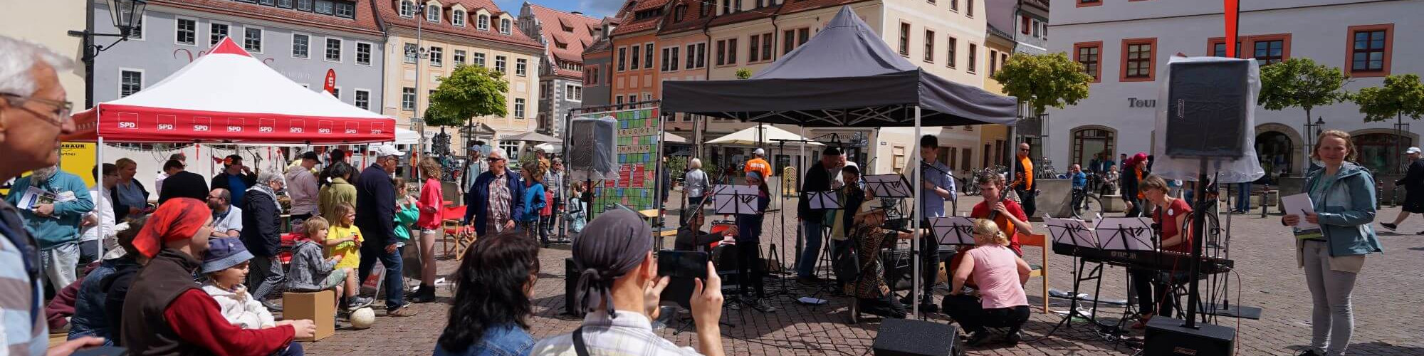 KI generiert: Das Bild zeigt eine belebte Straßenveranstaltung mit Menschen, die sich um Stände und eine kleine Bühne mit Musikern versammeln. Der Hauptinhalt ist ein öffentliches Fest oder Markt in einer historischen Stadtkulisse.