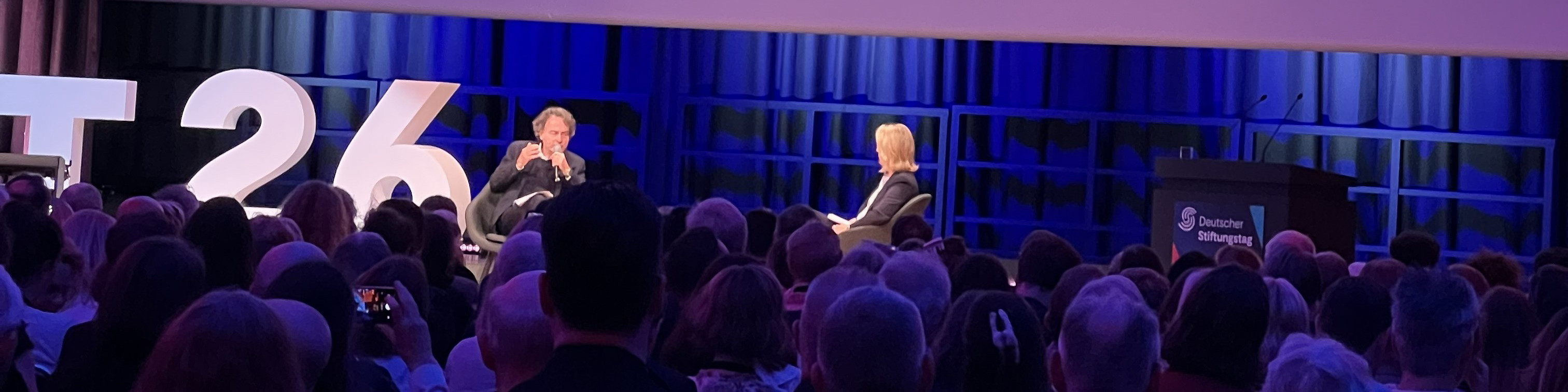 KI generiert: Das Bild zeigt eine Veranstaltung mit zwei Personen auf einer Bühne, die vor einem Publikum sprechen. Im Hintergrund sind eine große Zahl "26" und ein Podium mit einem Mikrofon zu sehen.