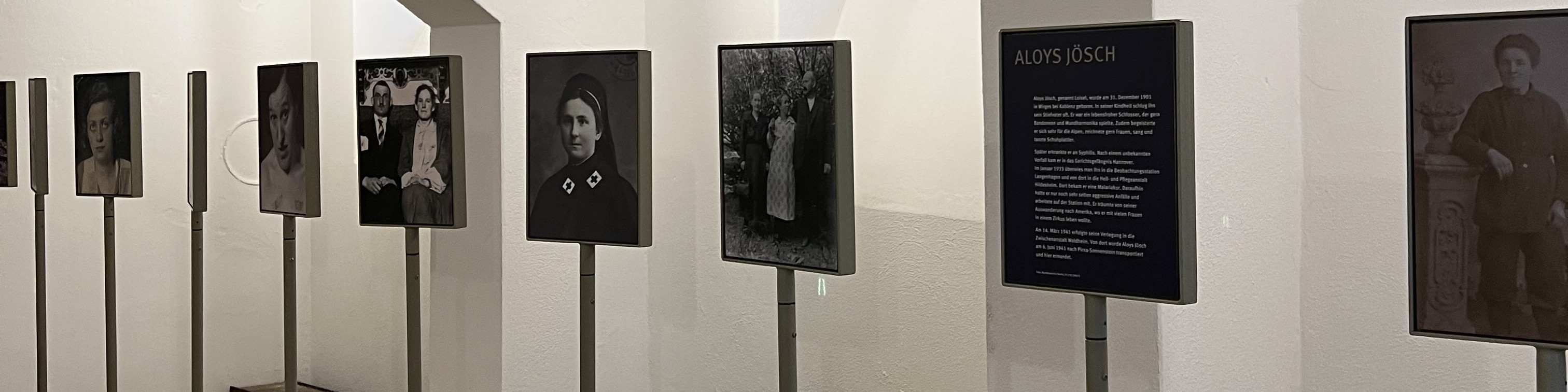 KI generiert: Das Bild zeigt eine Ausstellung mit mehreren schwarz-weißen Fotografien, die an Ständern befestigt sind. Der Hauptinhalt der Ausstellung scheint die Präsentation historischer Porträtfotografien zu sein, begleitet von einer Informationsafel mit dem Titel "Alois Jösch".