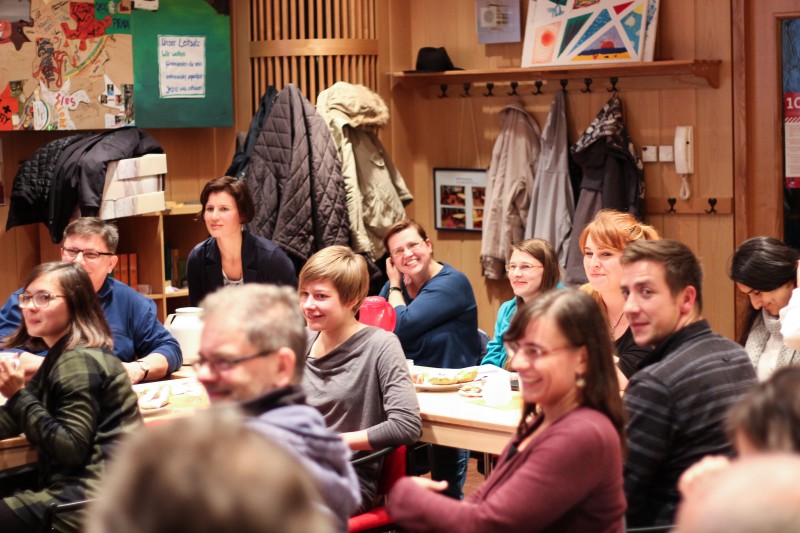 KI generiert: Das Bild zeigt eine Gruppe von Menschen, die in einem Raum mit Holzvertäfelung und Jacken an der Wand sitzen und sich unterhalten. Der Hauptinhalt des Bildes ist eine gesellige Zusammenkunft oder ein Treffen in einem gemütlichen, informellen Umfeld.