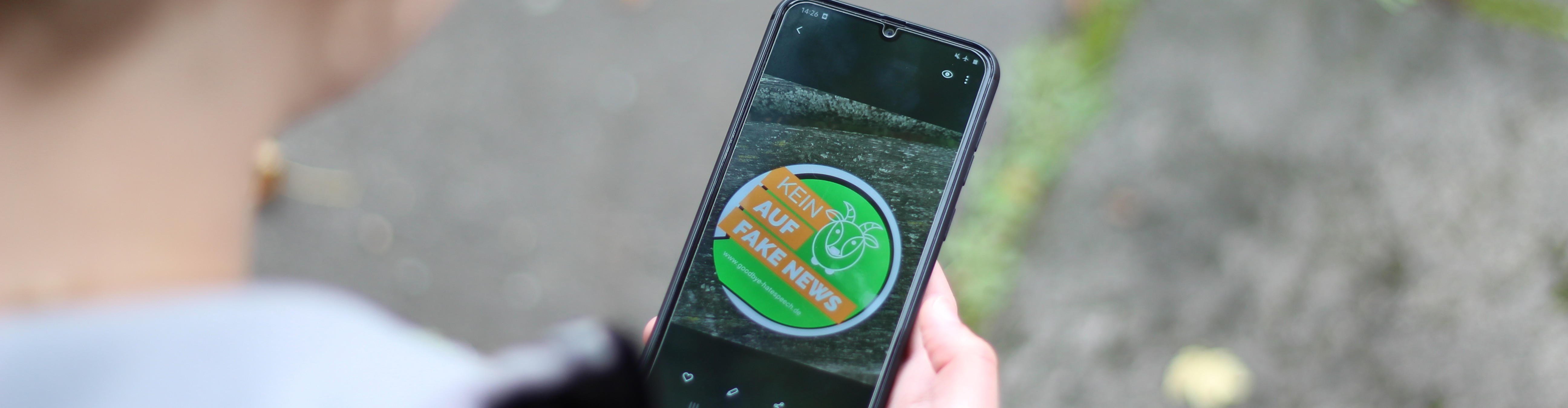 KI generiert: Eine Person hält ein Smartphone, auf dem ein Aufkleber mit der Aufschrift "Kein Platz für Fake News" zu sehen ist. Der Aufkleber ist grün und orange gestaltet.