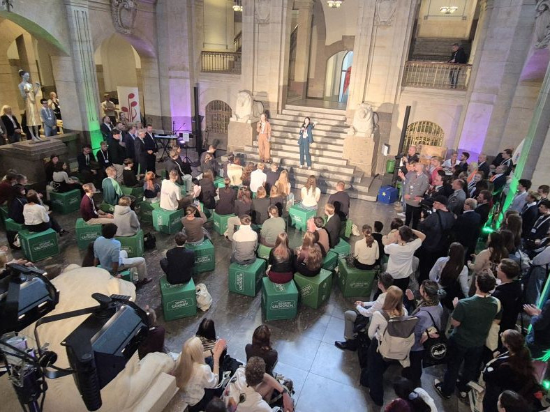 KI generiert: Das Bild zeigt eine große Menschenmenge in einem historischen Gebäude mit Säulen und einer Treppe, die an einer Veranstaltung teilnimmt. Im Vordergrund sitzen viele Personen auf grünen Sitzkissen, während im Hintergrund eine Bühne mit Moderatoren und Gästen zu sehen ist.